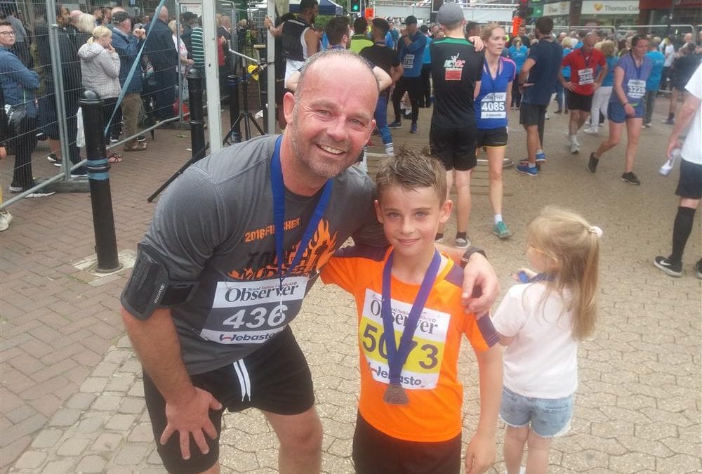 A man and a young boy wearing running clothes. They are wearing medals with purple ribbons. There is a large group of runners behind them.