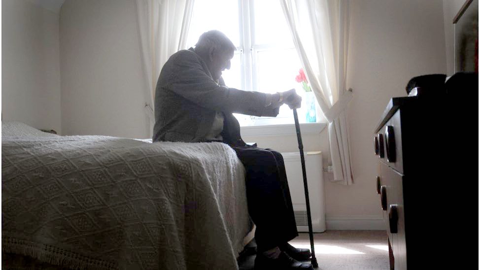 Man with walking stick sat on a bed. The man looks lonely.