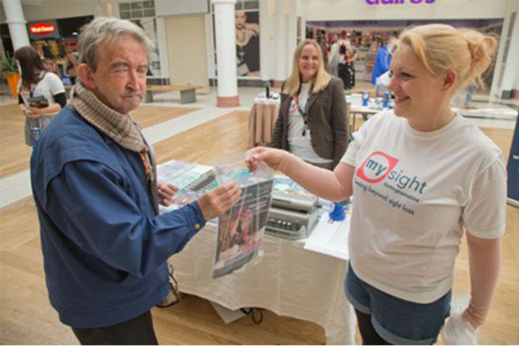 Woman wearing a My Sight Notts t-shirt handing a man information at a stall in the Victoria Centre Nottingham.