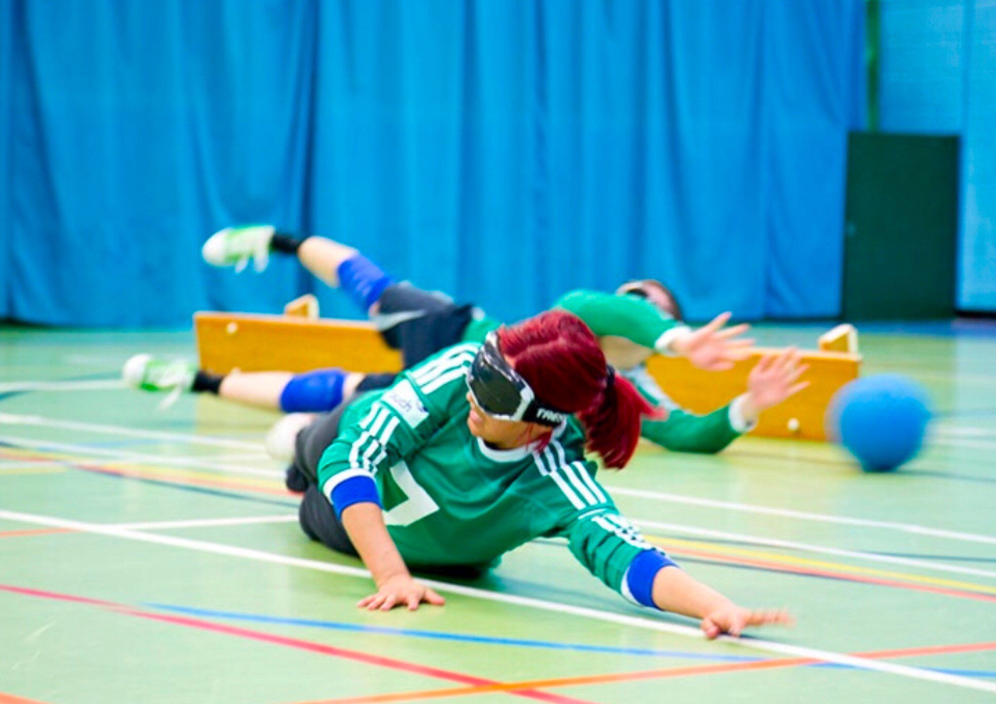 A woman laying on the floor of a Goal-ball court reaching for the ball. There is a man reaching for the ball behind her.