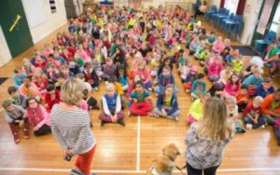 two women, one is a teacher and the other has a guide dog. They are stood in front of a large group of children in. an assembly hall in a school.