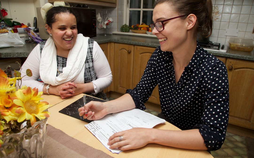 Two woman sat at a table in a kitchen chatting while filling out a form.