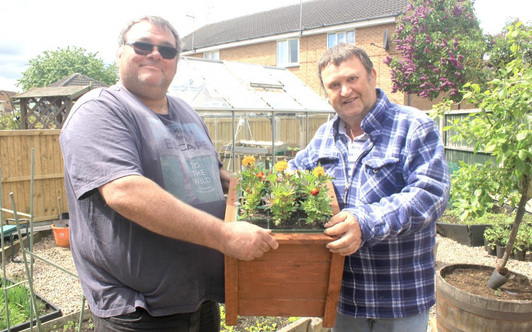 Two men holing a planter filled with flowers. they are in a garden with a greenhouse.