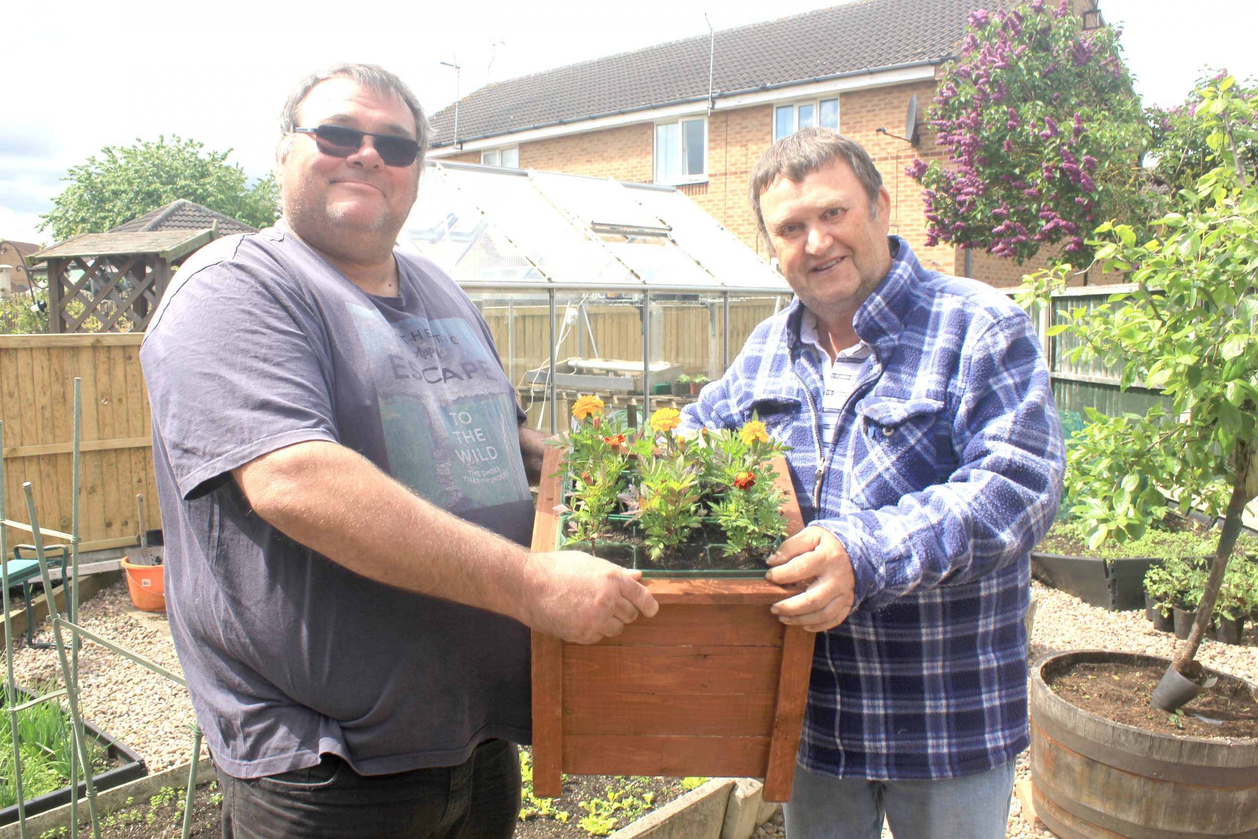 Two men holing a planter filled with flowers. they are in a garden with a greenhouse.