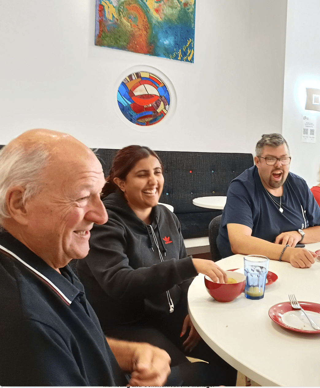 One woman and two men seated at a table laughing and eating crisps.