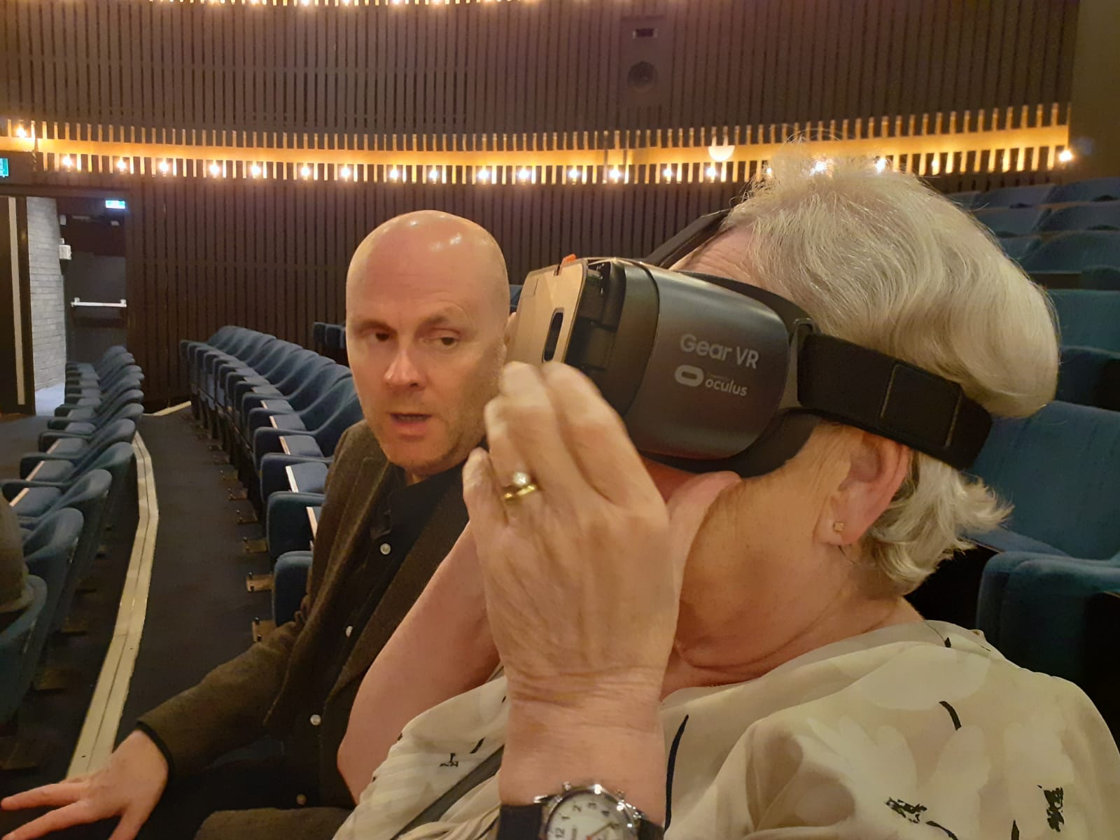 Photo shows Matt and Joan at the theatre. Joan is wearing a headset which she cups with her left hand as she focuses on the stage in front of her.