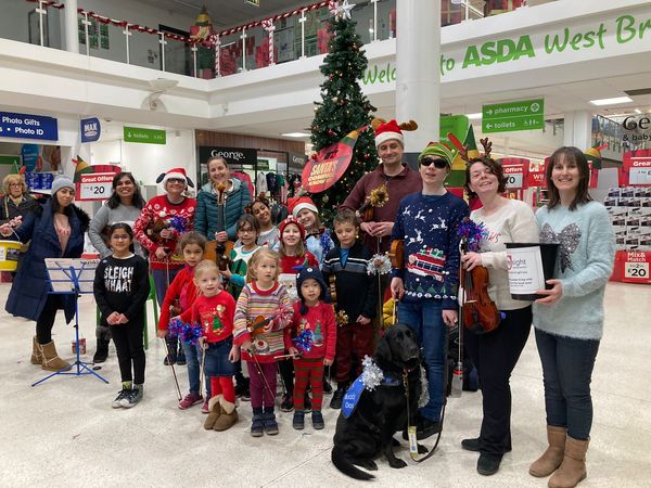 Seven adults and ten children all wearing Christmas seasonal jumpers and holding violins. There is also a guide dog in the foreground. They are located in ASDA West Bridgeford Nottingham.