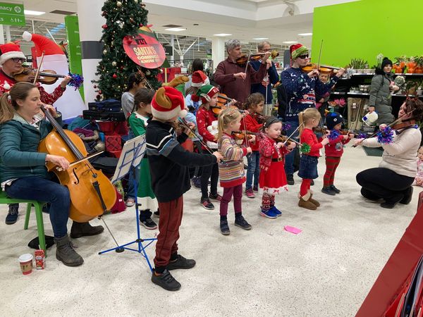 Seven adults and ten children all wearing Christmas seasonal jumpers and holding violins. They are located in ASDA West Bridgeford Nottingham.