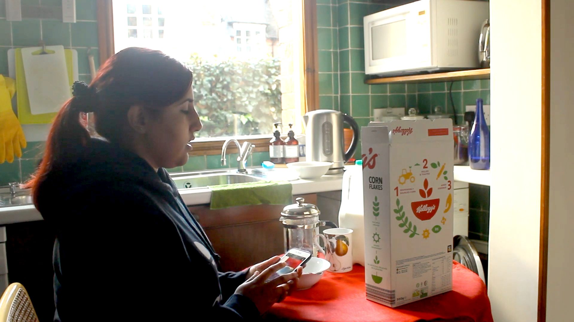 A woman in her kitchen using a mobile phone to scan a barcode to find out what the product in from of her is; which is cornflakes