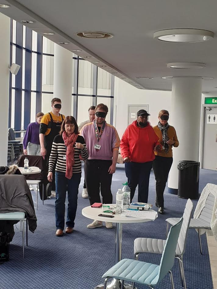 People in pairs, one learning to guide and the other wearing a blindfold and being guided around one of the venue’s foyer