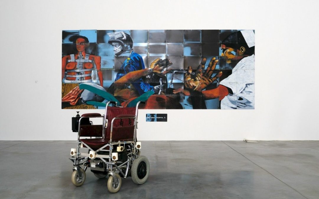 Picture shows an old red leather motorised wheelchair in front of a large painting in a gallery. On the left is an image of a woman in a metallic medical corset (after Mexican artist, Frida Kahlo). Across the bottom of the paining, the artist lies in a hospital bed holding his arms up in defence. A member of the Metropolitan Police’s infamous Special Patrol Group, with riot shield and helmet hovers over the artist. A nurse tends to the patient, attempting to pacify him.